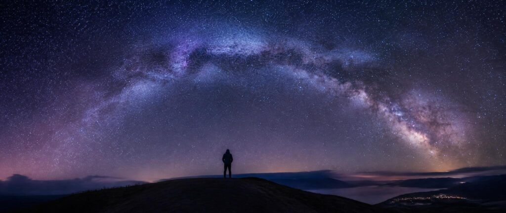 A small lone figure gazing up at the vast Milky Way, evoking a grounded version of the Overview Effect